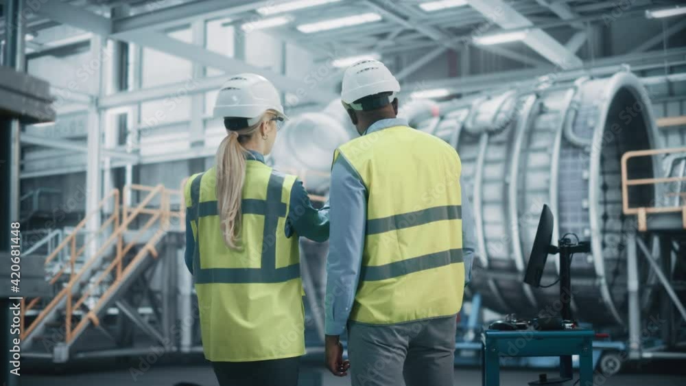 Two Diverse Professional Heavy Industry Engineers Wearing Safety Uniform and Hard Hats Working on Laptop Computer. African American Technician and Female Worker Talking on a Meeting in a Factory.