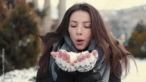 Winter girl portrait. Beauty joyful model girl blowing snow, having fun in winter park. Beautiful young woman enjoying nature outdoors. Wintertime.