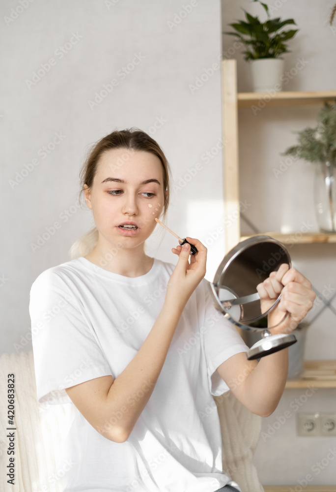 a young teenage woman in a white T-shirt masks pimples on her face with a foundation in front of a mirror. Care for sick skin of the face. Red spots on the skin. Skin problems in transition age a young teenage woman in a white T-shirt masks pimples on her face with a foundation in front of a mirror. Care for sick skin of the face. Red spots on the skin. Skin problems in transition age