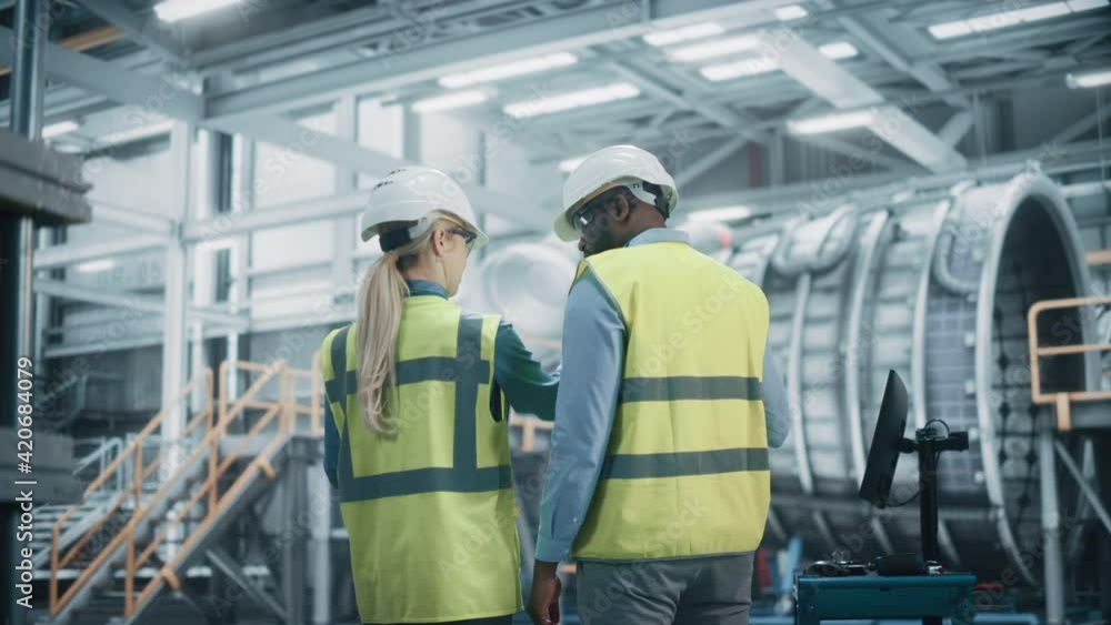 Two Diverse Professional Heavy Industry Engineers Wearing Safety Uniform and Hard Hats Working on Laptop Computer. African American Technician and Female Worker Talking on a Meeting in a Factory. Two Diverse Professional Heavy Industry Engineers Wearing Safety Uniform and Hard Hats Working on Laptop Computer. African American Technician and Female Worker Talking on a Meeting in a Factory.