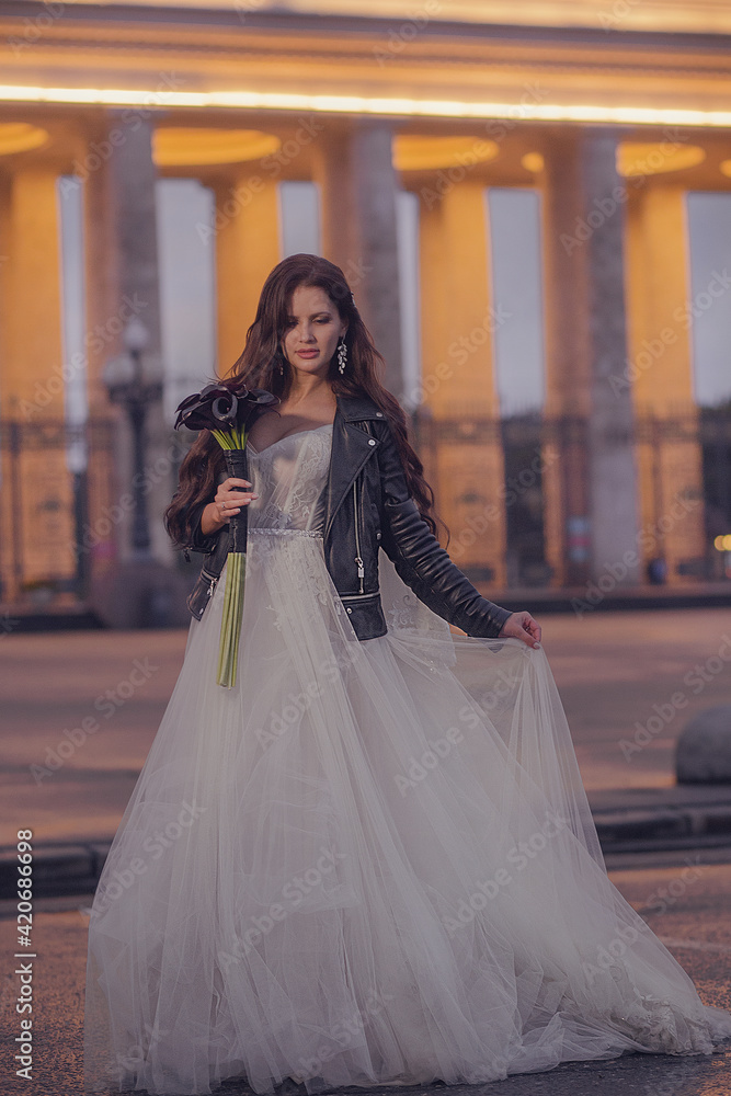 Naklejka premium A beautiful bride in a wedding dress and a leather jacket with a bouquet of flowers. Wedding concept. Evening photography