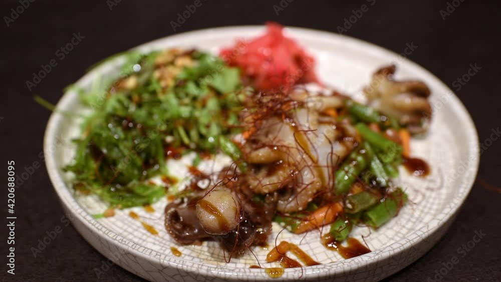 A moving video of the white ceramic plate with ginger, roasted vegetables, and octopuses pouring with soy sauce and sesame. A moving video of the white ceramic plate with ginger, roasted vegetables, and octopuses pouring with soy sauce and sesame.