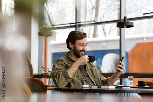 Unshaven happy man in earphones using mobile phone while drinking coffee