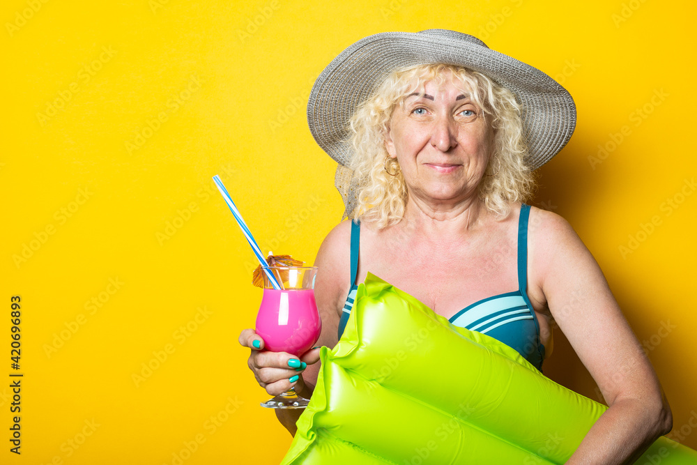 Old woman in swimsuit with cocktail holds air mattress on yellow background.