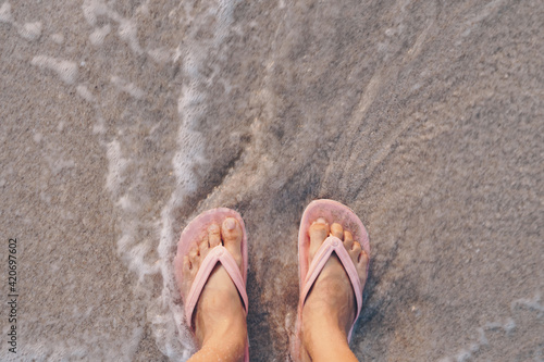 Woman feet wear slippers stand on sand tropical beach with sea water splash.