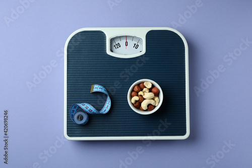 Scales with measuring tape and bowl of nuts on violet background
