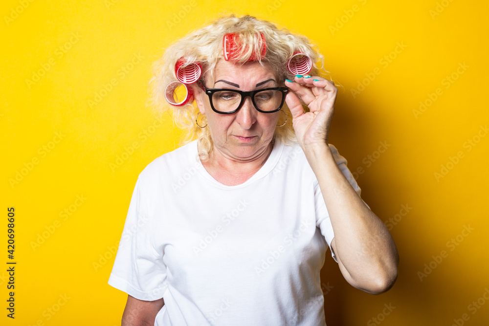 Pensive old woman with curlers in glasses looks down on a yellow background Pensive old woman with curlers in glasses looks down on a yellow background