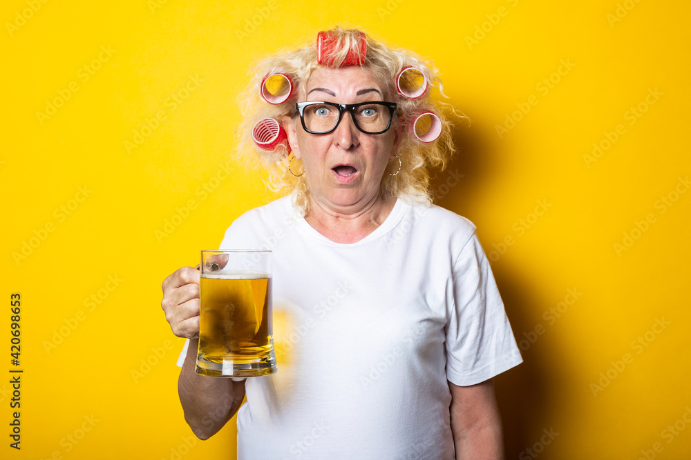 Surprised old woman with hair curlers with a glass of beer on a yellow background Surprised old woman with hair curlers with a glass of beer on a yellow background