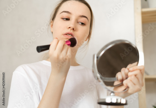novice makeup artist - a young teenage woman in a white T-shirt does makeup at home on her own in front of a mirror, applying a cosmetic toning agent to the skin of the face with a brush.