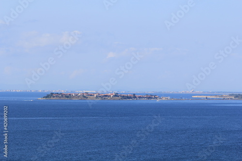 In the middle of the blue sea there is a peninsula with low old buildings and the other shore on the horizon