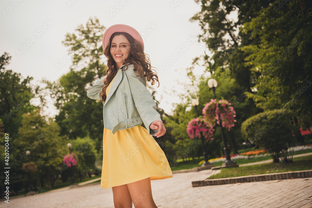 Photo of cute shiny young woman dressed teal jacket cap glasses walking smiling outdoors city park