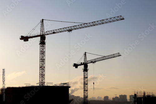 Silhouettes of construction cranes and unfinished residential building against the sunrise above the city. Housing construction, apartment block
