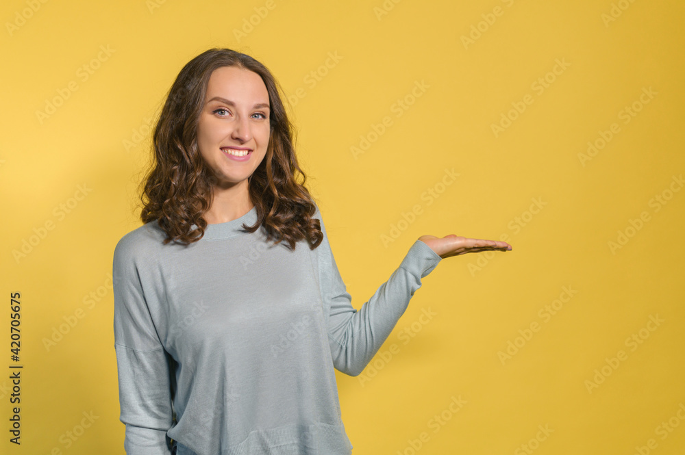 Cheerful curly young woman points hand at empty copy space, holding something on the palm of hand, mockup, smiling student girl presenting novelty, isolated on yellow Cheerful curly young woman points hand at empty copy space, holding something on the palm of hand, mockup, smiling student girl presenting novelty, isolated on yellow