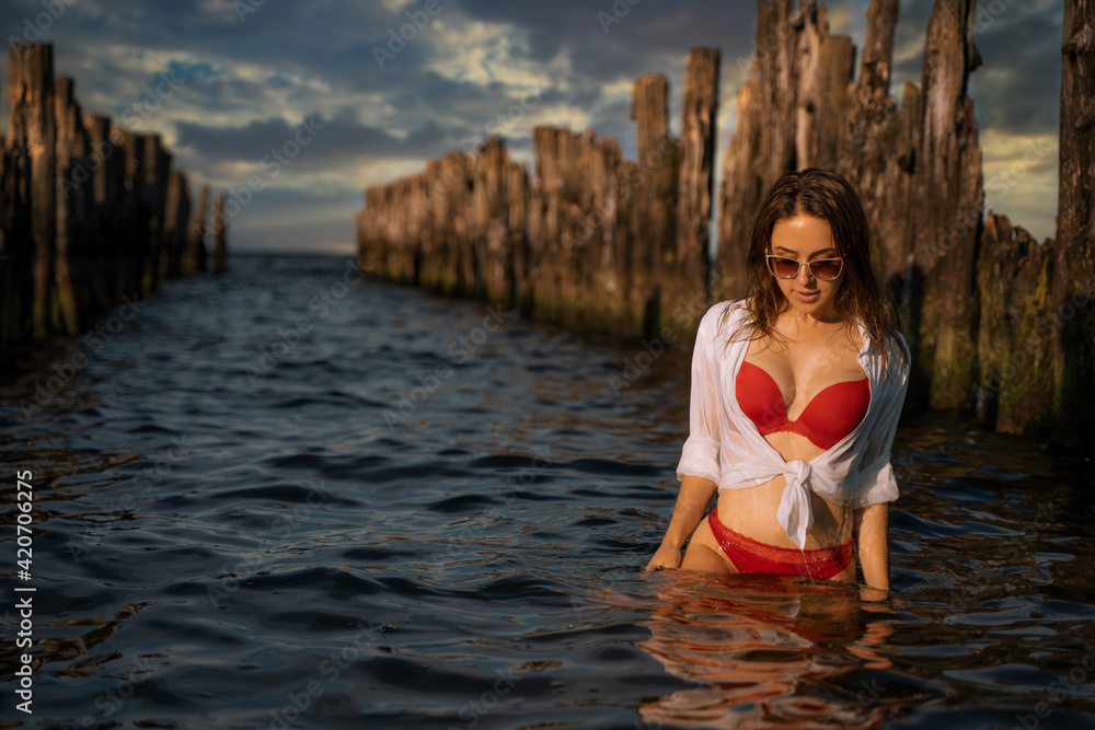 Fototapeta premium Young beautiful female brunette model pose in her red swimwear lingerie and white shirt over in the water between old pier wood bulks during dramatic sunset