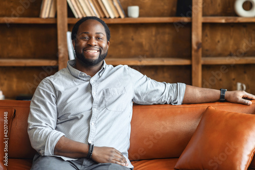 Ethnic black unshaved plump man with dreadlocks sitting on the leather sofa and smiling, in a normal blue shirt and jeans, feeling comfort and relaxation, at an interview, bookcase in the background