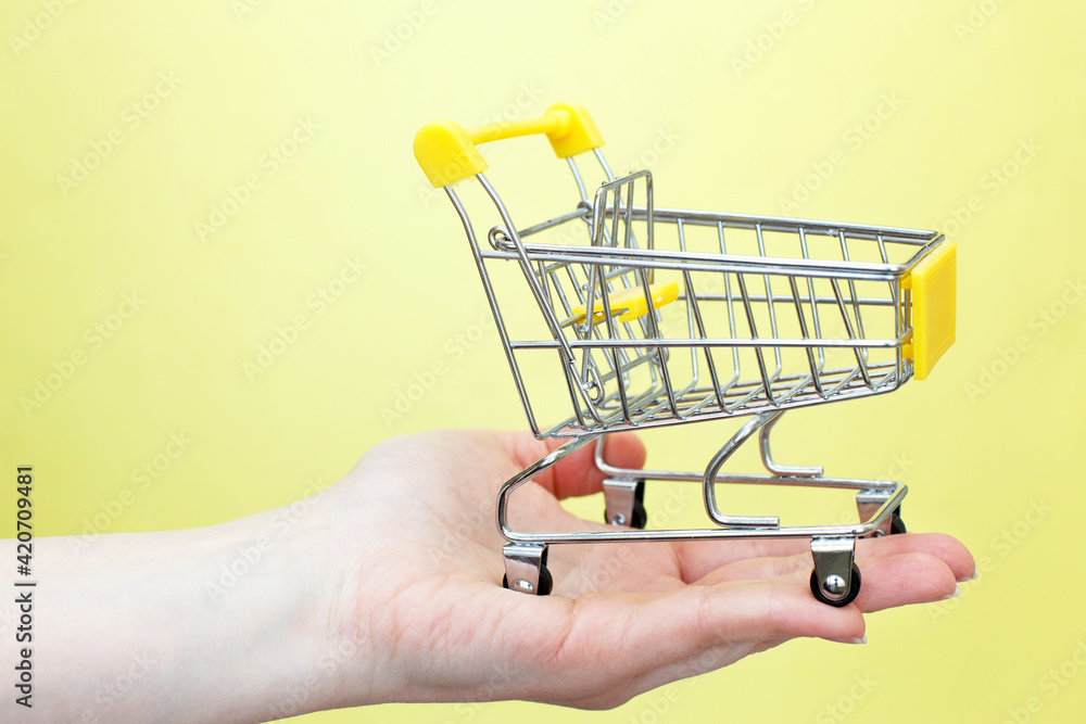 The shopping cart in female hand on yellow background The shopping cart in female hand on yellow background