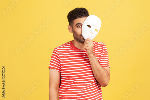 Funny crazy man with beard in striped t-shirt peeping out white face mask, hiding his real feelings, pretending to be another person. Indoor studio shot isolated on yellow background photo