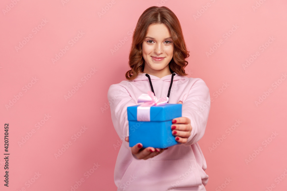 Portrait of cheerful brunette teenage girl in hoodie presenting blue wrapped gift box at camera and sincerely smiling, birthday greetings. Indoor studio shot isolated on pink background Portrait of cheerful brunette teenage girl in hoodie presenting blue wrapped gift box at camera and sincerely smiling, birthday greetings. Indoor studio shot isolated on pink background