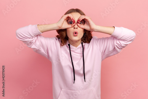 Portrait of amazed teen girl with curly hair in hoodie looking through binoculars gesture and expressing surprise, zooming vision, exploring distance. Indoor studio shot isolated on pink background © khosrork