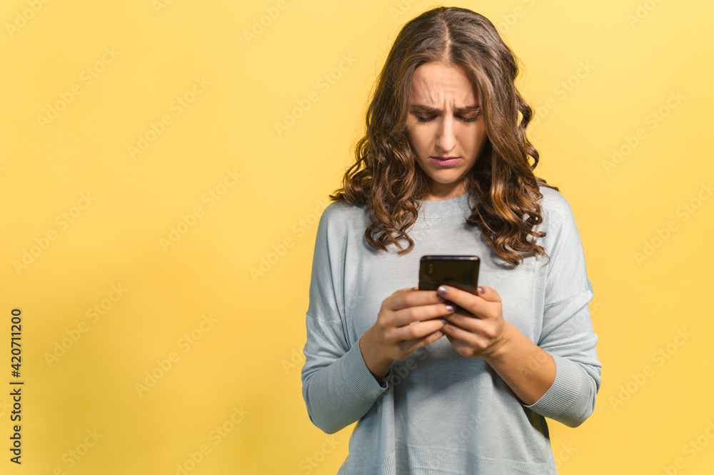 Upset curly brunette young woman holding smartphone, looking with disappointment face expression at phone screen, received a bad comment in social networks from hater, dislike, isolated on yellow Upset curly brunette young woman holding smartphone, looking with disappointment face expression at phone screen, received a bad comment in social networks from hater, dislike, isolated on yellow