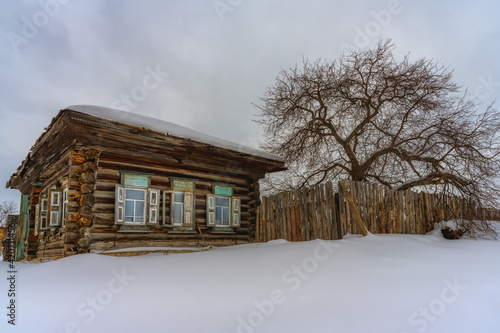 An old log village house with a fence and a tall tree nearby on a cloudy winter day. Windows with carved frames, high snowdrifts. Kashina village (Bogdanovichi district, Sverdlovsk region, Russia)