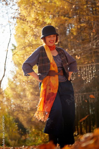 Mature woman in interesting dress and long orange scarf in autumn park and trees with yellow leaves background