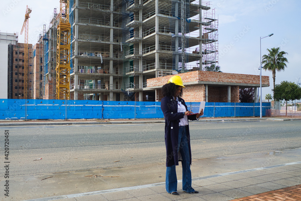 African-American black woman construction manager in yellow hardhat set out to check the construction of the flats. She is holding a computer in her hand to check the progress of the construction