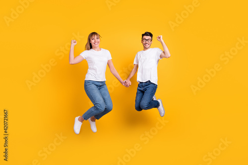 Full length photo of adorable sweet persons dressed white t-shirt jumping holding hands rising fists isolated yellow color background