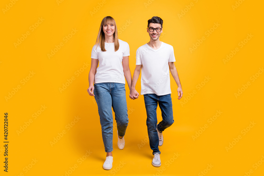 Full length photo of funky cute two persons dressed white t-shirt walking holding hands arms isolated yellow color background Full length photo of funky cute two persons dressed white t-shirt walking holding hands arms isolated yellow color background