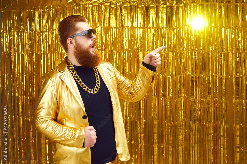 Surprised chubby man in extravagant shiny golden disco jacket, gold chain  necklace and cool glasses pointing index finger aside to advertising copy  space. Show, event, party, concert ad background Stock Photo