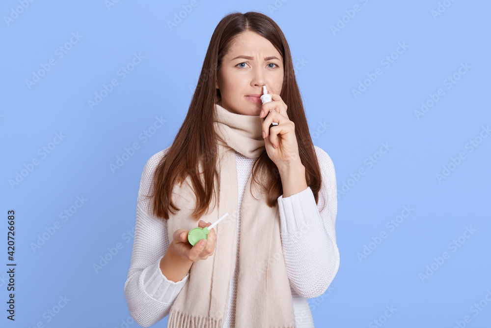 Woman using nasal spray and looking directly at camera with frowning face. Lady catching cold and need treatment, dark haired girl wearing casual jumper and scarf feels unwell. Woman using nasal spray and looking directly at camera with frowning face. Lady catching cold and need treatment, dark haired girl wearing casual jumper and scarf feels unwell.
