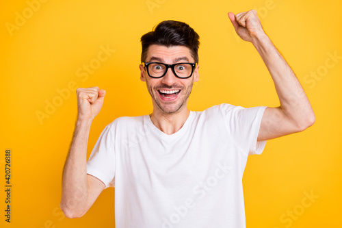 Photo of funny lucky young man wear white t-shirt spectacles rising fists isolated yellow color background © deagreez