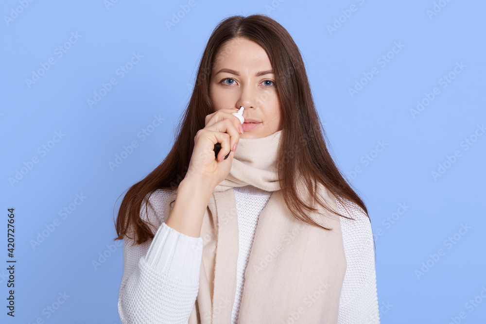 Young dark haired woman wearing white sweater and scarf standing dripping nasal drops isolated on blue background, ill lady looks at camera with upset facial expression. Young dark haired woman wearing white sweater and scarf standing dripping nasal drops isolated on blue background, ill lady looks at camera with upset facial expression.