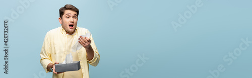 man sneezing and holding tissue box with napkins isolated on blue, banner