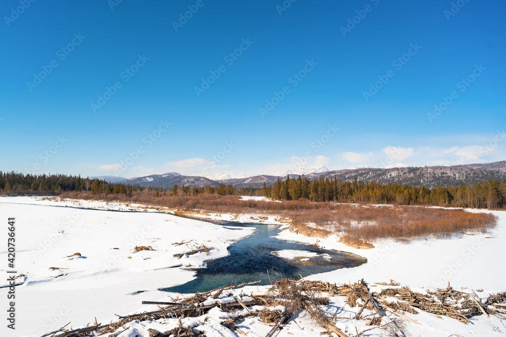 Turquoise water flows among the white snows against the backdrop of the mountains. This is the Irkut River in the reserved Tunkinskaya Valley on a sunny day in early spring. 