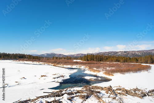 Turquoise water flows among the white snows against the backdrop of the mountains. This is the Irkut River in the reserved Tunkinskaya Valley on a sunny day in early spring. 