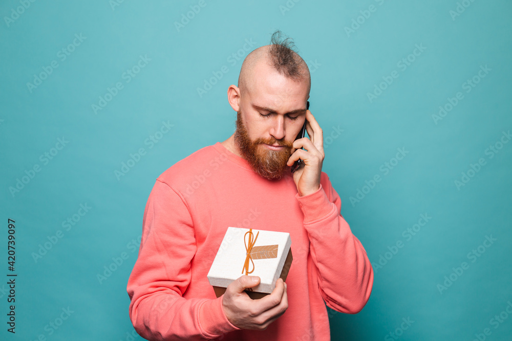 Bearded european man in casual peach isolated on turquoise background holding gift box talk on mobile phone with serious face Bearded european man in casual peach isolated on turquoise background holding gift box talk on mobile phone with serious face