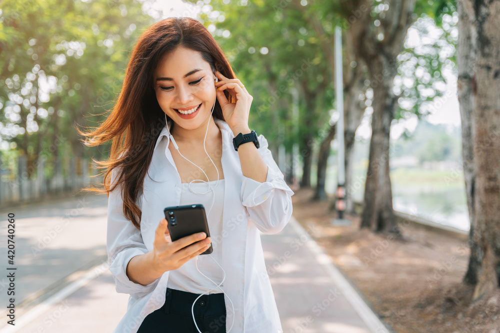 Asian woman using the small talk headphones to make a happy phone call in the park. Asian woman using the small talk headphones to make a happy phone call in the park.