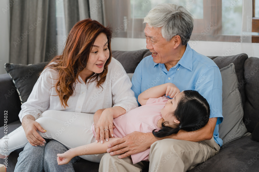 Obraz premium Asian grandparents talking with granddaughter at home. Senior Chinese, old generation, grandfather and grandmother using family time relax with young girl kid lying on sofa in living room concept.
