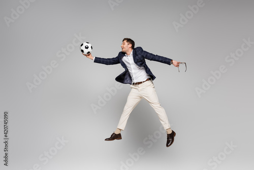 full length of smiling businessman holding glasses and playing football while levitating on grey