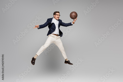 full length of cheerful man in glasses playing basketball and levitating on grey