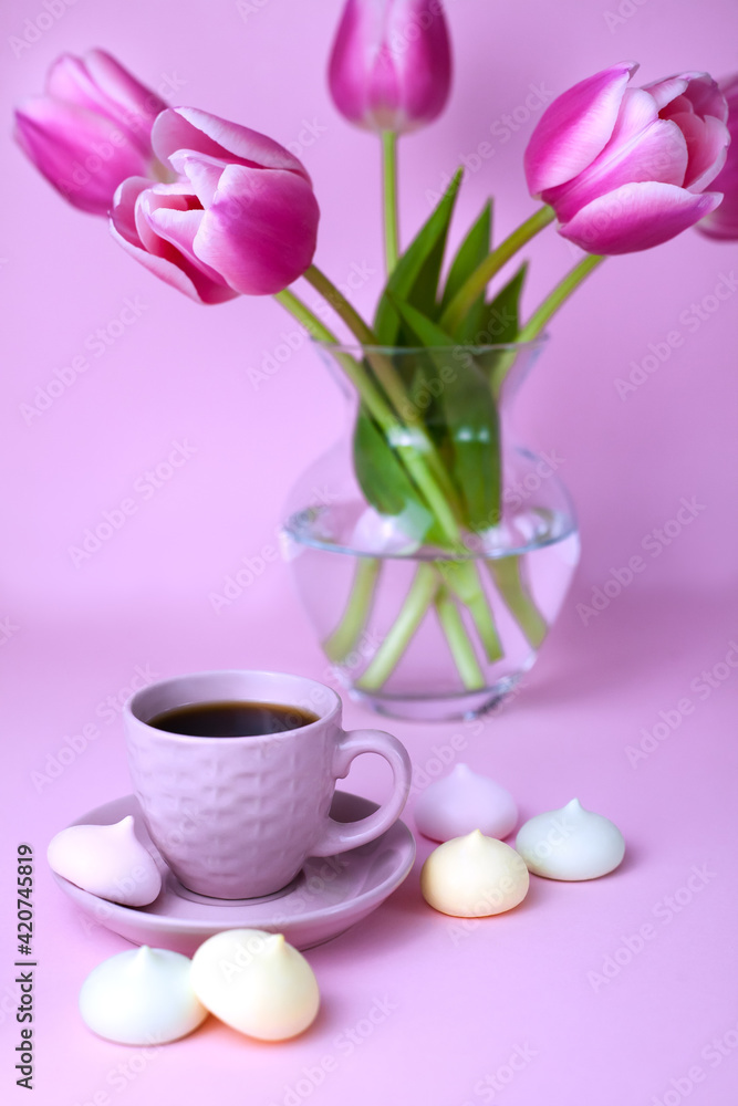 A cup of fresh coffee and a glass vase with pink tulips on the pink background. Close-up. A cup of fresh coffee and a glass vase with pink tulips on the pink background. Close-up.