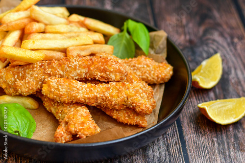  Crispy deep fried chicken strips with sesame seeds and french fries . Breaded with cornflakes chicken breast fillets with chilly peppers and fresh basil on wooden rustic background