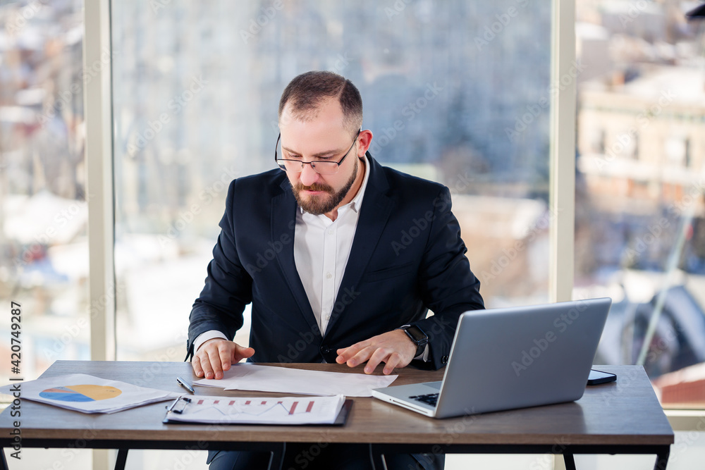 Adult male mentor, director, businessman in glasses and a suit studying documents while sitting at the table. Working day concept