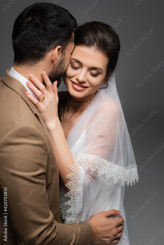 smiling bride with closed eyes hugging muslim fiance isolated on grey