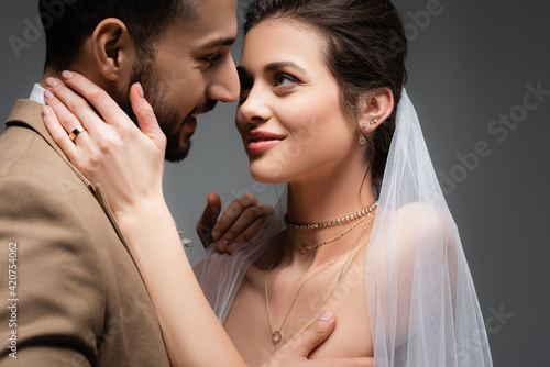elegant woman in veil embracing neck of muslim groom isolated on grey