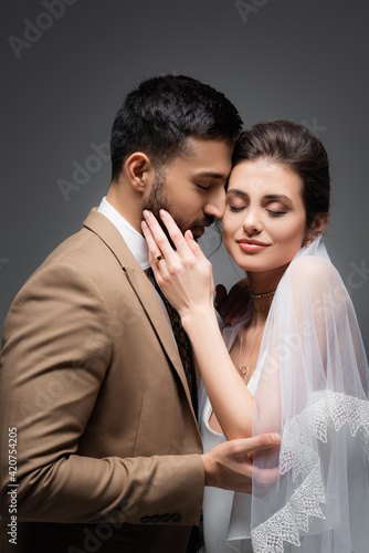 smiling bride with closed eyes touching face of elegant muslim man isolated on grey