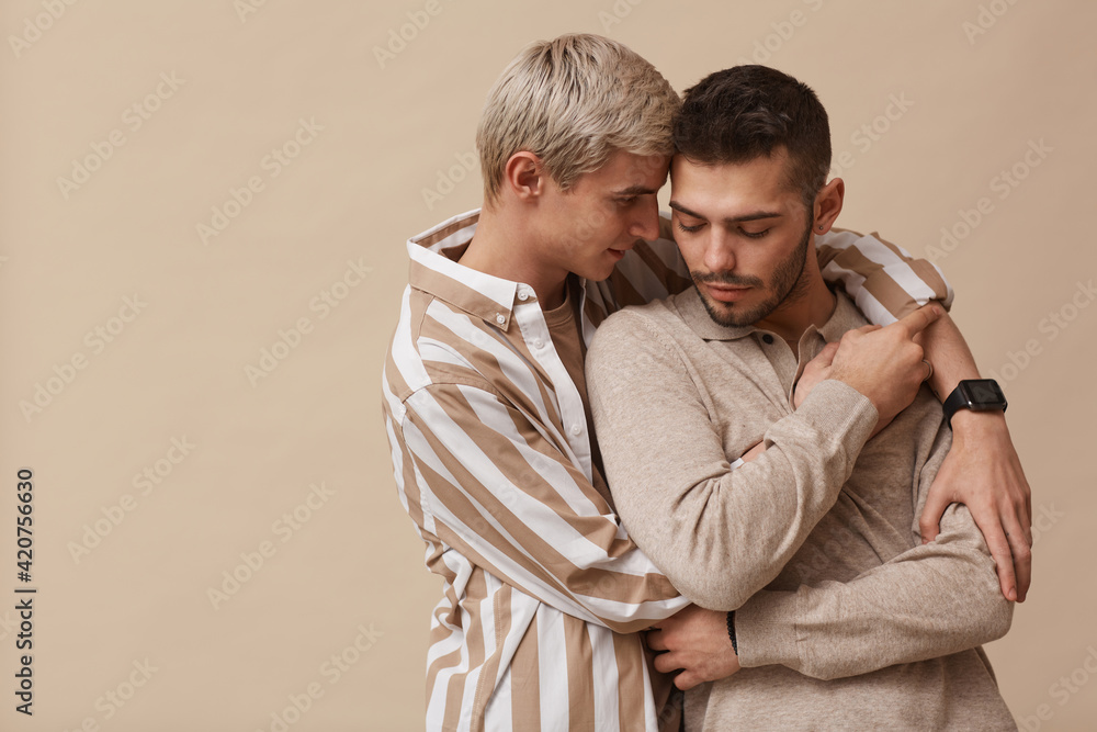Minimal waist up portrait of loving gay couple embracing while posing against neutral beige background in studio, copy space