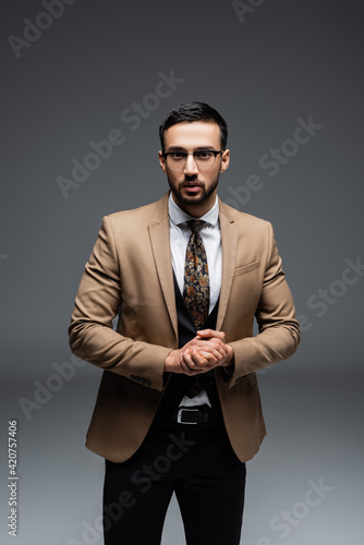 muslim man in stylish suit and eyeglasses standing with clenched hands on grey