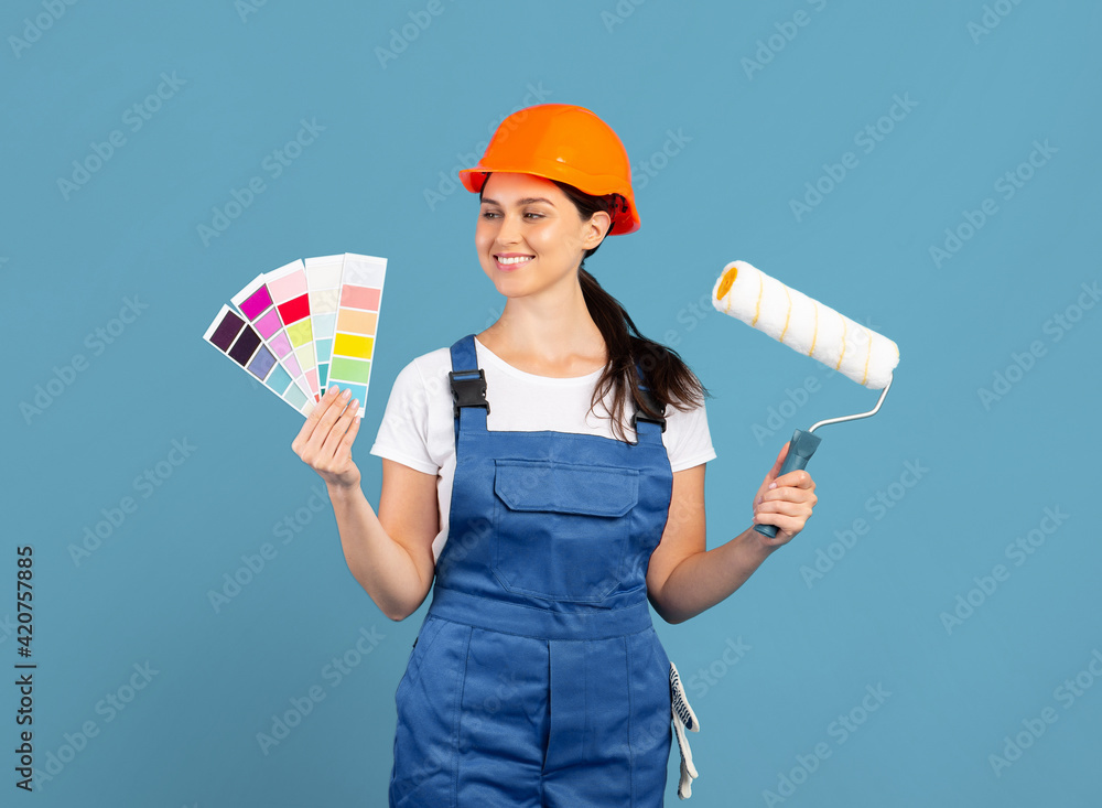 Smiling handywoman in coveralls and hardhat holding color palette and roller brush